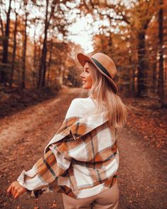 a woman is walking down a dirt road in the woods wearing a plaid shirt and hat