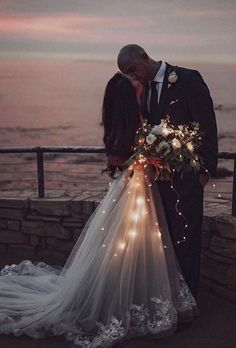 a bride and groom kissing in front of the ocean at night with lights on their veil