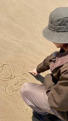 a little boy sitting on the beach writing in the sand with his hat over his head