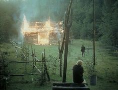 a person sitting on a bench in front of a log cabin with flames coming from it