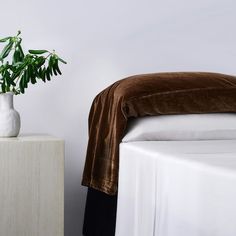 a white vase with green plants in it next to a brown blanket on a bed