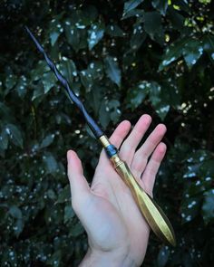 a hand holding a gold colored knife in front of some bushes and trees with green leaves