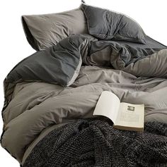 an open book sitting on top of a bed with gray sheets and pillows, next to a black blanket