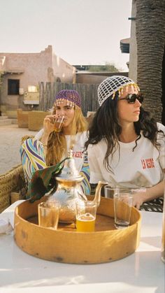 two women sitting at an outdoor table with drinks in front of them, one drinking from a teapot