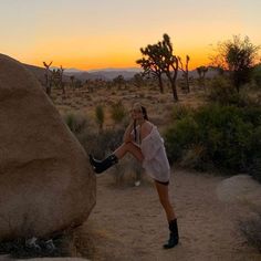 a woman standing next to a large rock in the middle of a dirt field at sunset