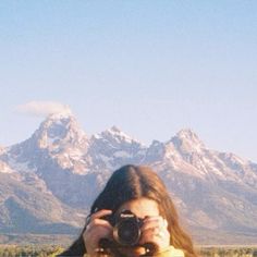 a woman holding a camera up to her face in front of mountains and snow capped peaks