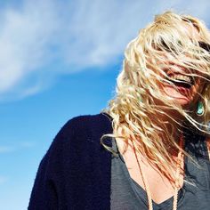 a blonde woman with her hair blowing in the wind and blue sky behind her is looking at the camera