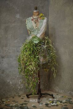 a mannequin is covered with plants and leaves in front of a concrete wall