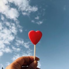 a hand holding a red heart shaped lollipop against a blue sky with clouds