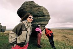 two women standing in front of a large rock on top of a grass covered field