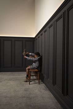 a man sitting on top of a wooden chair next to a tall black paneled wall