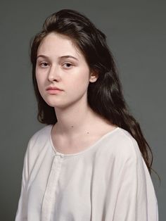 a woman with long dark hair wearing a white shirt and standing in front of a gray background