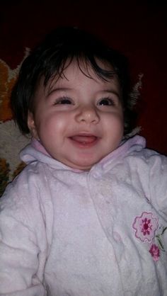 a smiling baby wearing a white jacket and pink flowered shirt on top of a bed