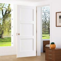 an open door in a white room next to a dresser and window with trees outside