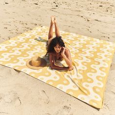 a woman laying on top of a yellow and white blanket next to the ocean while holding a hat