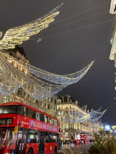 a red double decker bus driving down a street next to tall buildings covered in christmas lights