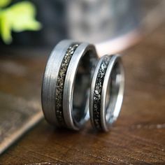 two wedding bands with black diamonds on them sitting on top of a wooden table in front of a plant