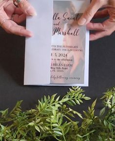 two hands holding up a wedding program with greenery in the foreground and on the background