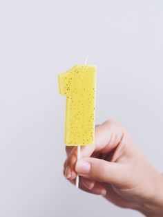 a hand holding a small yellow cake with a one on it's stick in front of a white background