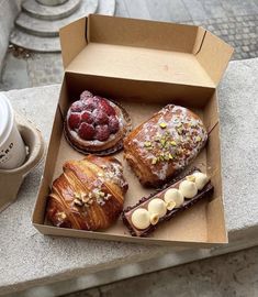 three pastries in a box next to a cup of coffee on a table outside