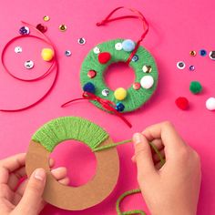 someone making a christmas wreath out of yarn and buttons on a pink surface with other crafting supplies