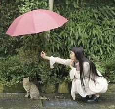 a woman kneeling down with an umbrella over her head and a cat on the ground