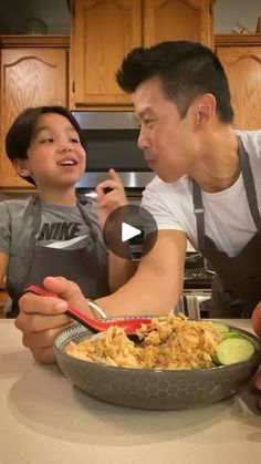 a man and boy sitting at a kitchen counter eating food from a bowl with spoons