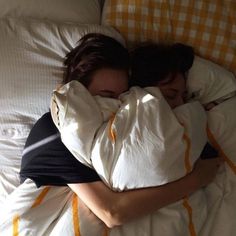 two people laying in bed with pillows and blankets on them, one is hugging the other