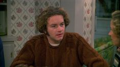 a man with curly hair sitting at a table talking to another person in the kitchen