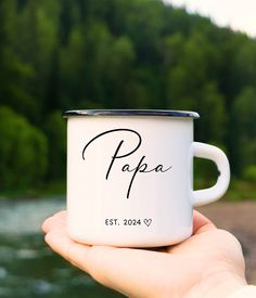 a person holding a white coffee mug with the word papa on it in front of a river