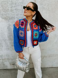 a woman in white pants and a blue jacket holding a silver handbag while standing next to a concrete wall