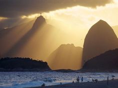 the sun shines through clouds over mountains and people on the beach in front of them