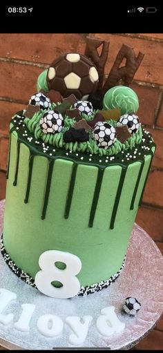 a green birthday cake with soccer balls and numbers on the bottom tier, sitting on top of a table next to a brick wall