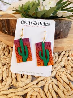 a pair of earrings with an orange and green cactus design on the front, sitting next to a potted plant