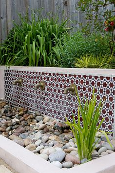 a garden with rocks, plants and water features on the side of the building that has a fenced in area next to it