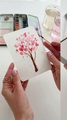 two pictures of someone holding up a card with a tree on it and hearts painted on it