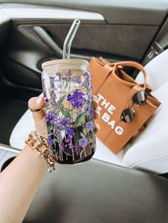 a woman holding a glass with purple flowers on it and a handbag in the background