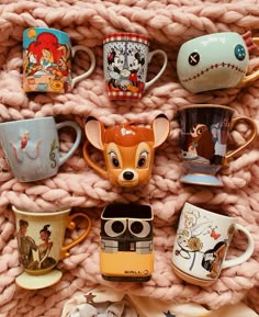 many coffee mugs are lined up together on a knitted blanket with the faces of cartoon characters