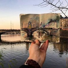 someone is holding up an open book in front of the water with buildings on it