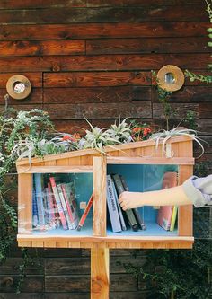 a person reaching for books in a wooden shelf with plants growing on the top and bottom