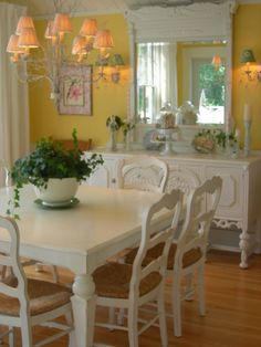 a white dining room table with chairs and a vase on top of it, in front of a yellow wall