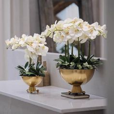 two gold vases with white flowers in them on a marble countertop next to a mirror