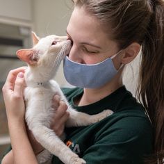 a woman wearing a face mask and holding a white cat in her arms while she holds it up to her mouth