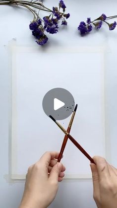 two hands holding pencils over a piece of paper with flowers on the table in the background