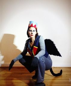 a woman dressed up as an evil queen with wings on her head and legs, crouching in front of a white wall