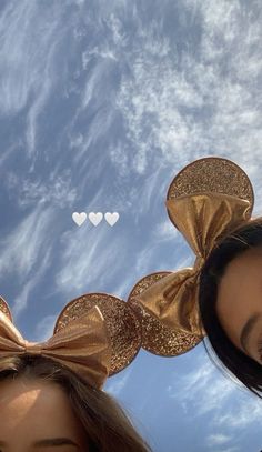 two people wearing gold minnie ears with the sky in the background and hearts drawn on them