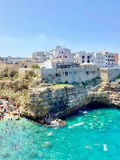 people are swimming in the blue water near an old stone cliff and buildings on the other side