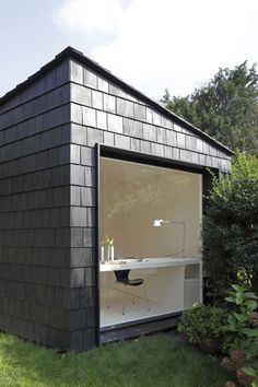 a small black building with a desk and chair in the front yard, surrounded by greenery