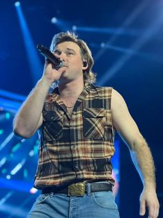 a male in a plaid shirt and jeans holding a microphone up to his mouth as he sings