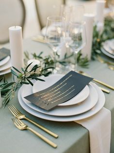 the table is set with white plates and silverware, gold utensils and greenery